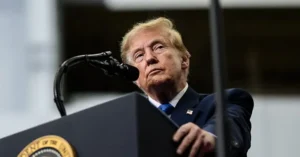 The image shows a person standing at a podium with a microphone, wearing a suit and tie, and appears to be addressing an audience.