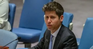 The image shows a man in a suit seated at a table with a microphone, likely participating in a formal meeting or conference.