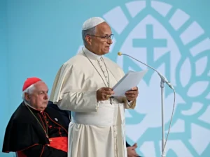 Pope Francis delivers a speech while holding a document, with a cardinal seated in the background.