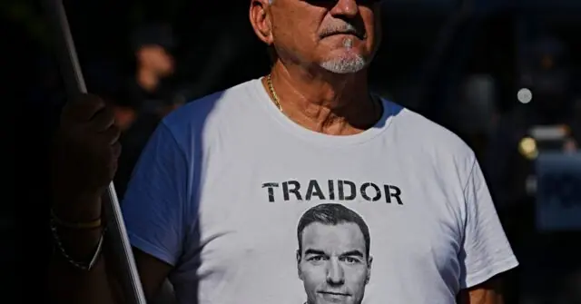 The photo shows a man wearing a T-shirt with a portrait and the word "TRAIDOR" printed on it, holding a flag.