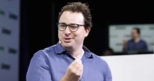 The image shows a man wearing glasses and a blue shirt, gesturing with his hand while speaking, likely during a presentation or speech.