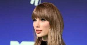 The image shows a person with long blonde hair and bangs, wearing a black top, against a blue background with a logo partially visible.