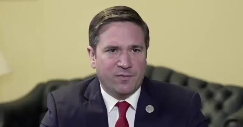 The image shows a man in a suit and red tie, seated in a formal setting, likely during a news interview or press conference.