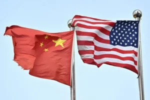 The photo shows the flags of China and the United States waving side by side against a clear sky, symbolizing diplomatic relations or international cooperation.