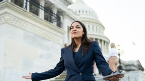 A woman in a suit stands in front of the U.S. Capitol, holding a folder and gesturing with her hands.