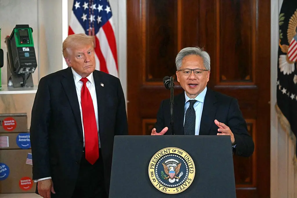 The image shows two individuals standing at a podium with the seal of the President of the United States, with one person speaking and the other standing beside them.