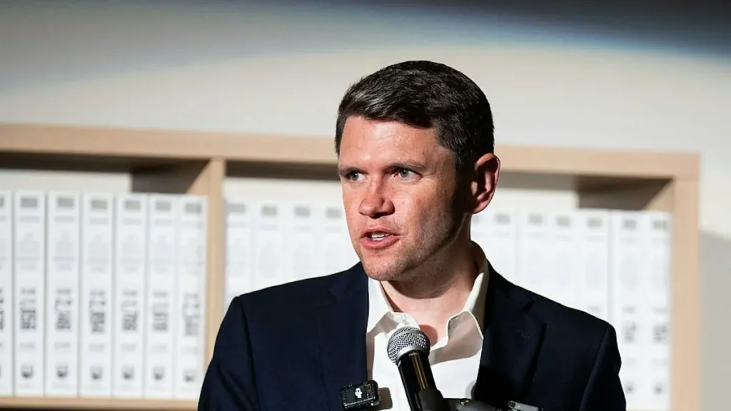 A man in a suit is speaking into a microphone in front of a shelf of binders.