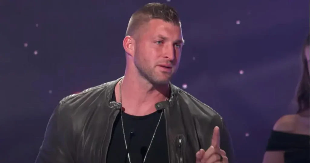 The image shows a man in a black leather jacket and a black shirt, gesturing with his right hand while speaking.
