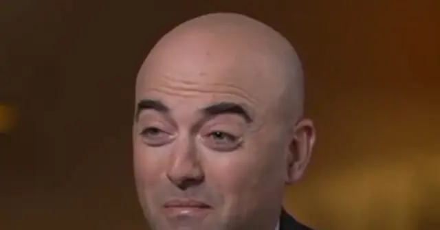 The image shows a close-up of a person with a bald head, wearing a suit, against a blurred background.