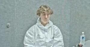 The image shows a person in a white jacket sitting in a courtroom setting.