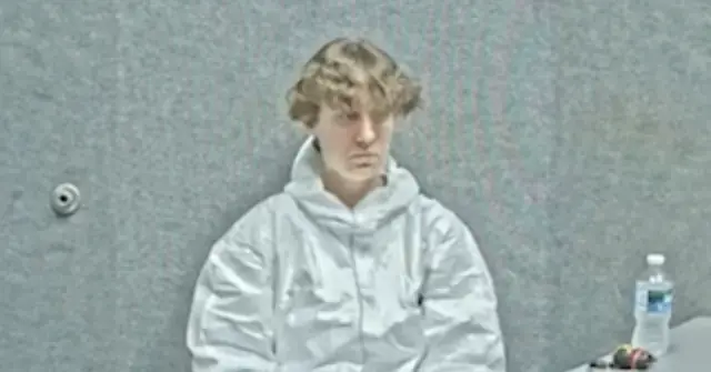The image shows a person in a white jacket sitting in a courtroom setting.