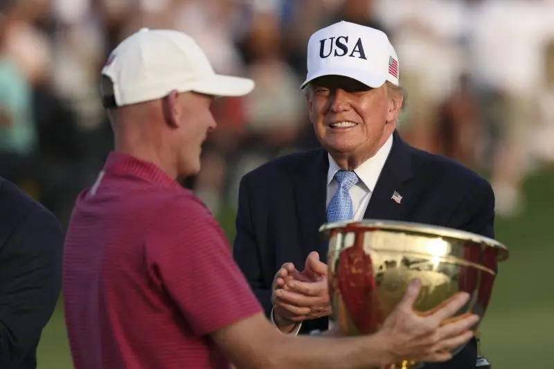 The image shows a person in a USA cap shaking hands with another individual while holding a large trophy, likely at a golfing event.