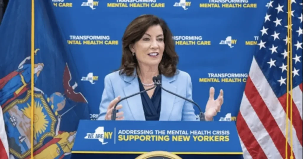 A woman speaks at a podium with a backdrop emphasizing mental health care, flanked by flags of New York and the United States.