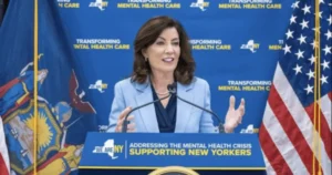 A woman speaks at a podium with a backdrop emphasizing mental health care, flanked by flags of New York and the United States.