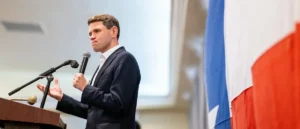 A man in a suit is giving a speech at a podium with a microphone, with a flag in the background.