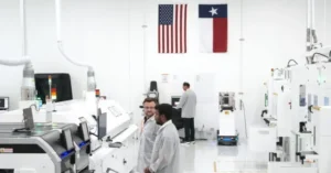 The image shows a cleanroom environment with individuals in lab coats working on advanced machinery, with American and Texas flags prominently displayed on the wall.