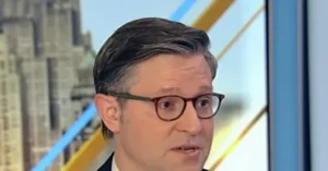 The image shows a man wearing glasses and a suit, appearing to be on a news broadcast with a cityscape in the background.