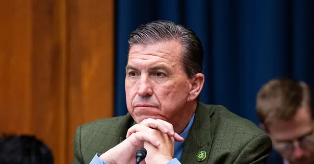 The image shows a man in a green suit with his hands clasped, appearing to be in a formal setting, possibly a hearing or meeting.