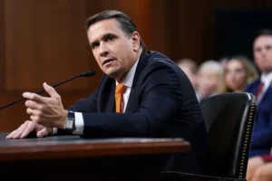A man in a suit and tie is speaking at a formal event, gesturing with his hand as he addresses the audience.