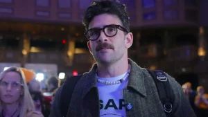 The image shows a man wearing glasses, a pearl necklace, and a "SAP" t-shirt, standing in a crowded urban setting at night.
