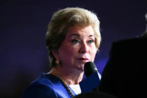 A woman in a blue suit is speaking into a microphone at a formal event.