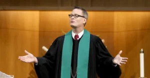A person in a black robe and green stole stands with arms outstretched in a church setting.
