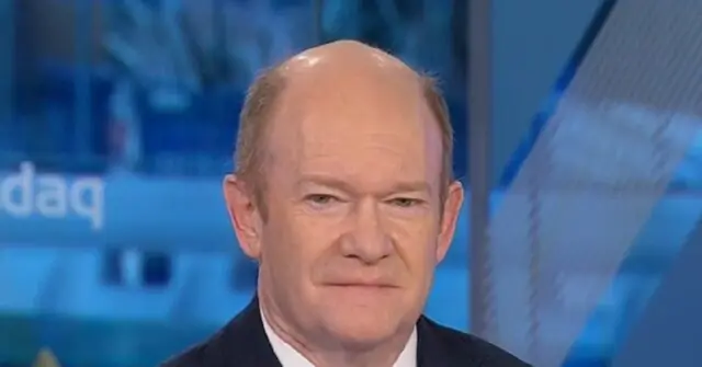 The image shows a man in a suit on a news broadcast, with a blurred background featuring the word "daq."