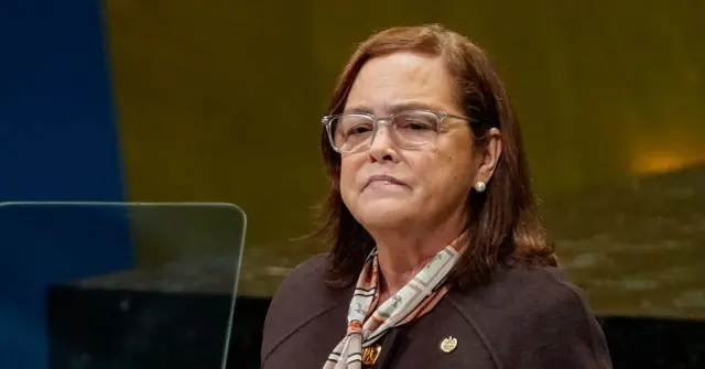 The image shows a person wearing glasses and a scarf, standing in front of a blue and yellow background, possibly during a formal or professional setting.