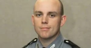 The image shows a person in a police uniform, likely a law enforcement officer, against a plain background.