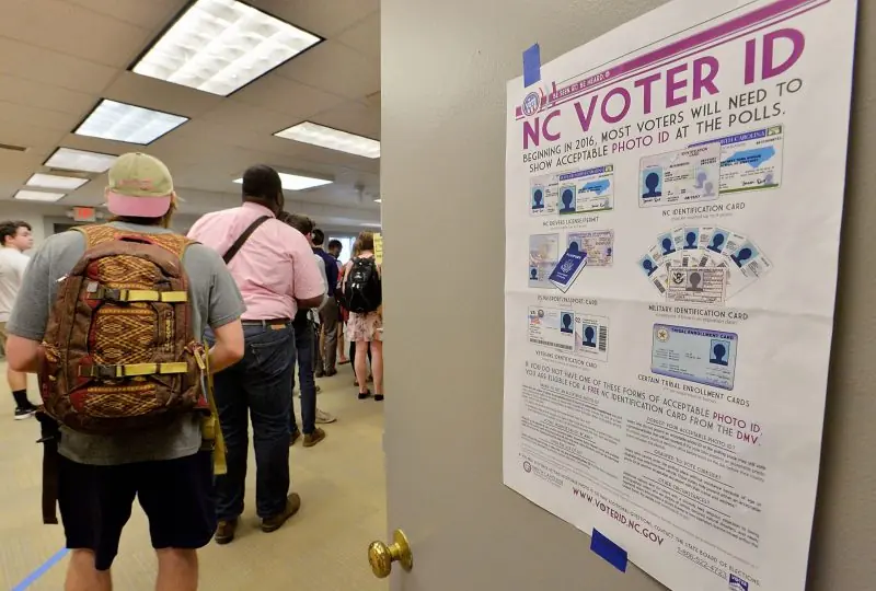 A poster about the NC Voter ID requirements is displayed as people wait in line, emphasizing the need for acceptable photo ID at the polls.