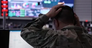 A soldier in a camouflage uniform is seen from behind, with hands on their head, in a setting that appears to be a control room with multiple screens displaying various data.