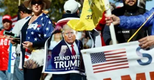 The photo depicts a group of people holding signs and banners supporting Donald Trump, including one that says "Stand With Trump."