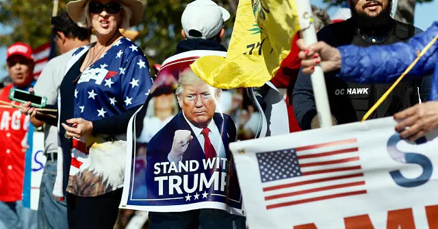 The photo depicts a group of people holding signs and banners supporting Donald Trump, including one that says "Stand With Trump."