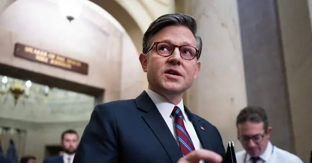 The image shows a man in a suit and tie speaking in what appears to be a formal or official setting, possibly a government or legislative building.