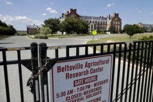 The image shows a locked gate with a sign for the Beltsville Agricultural Research Center, indicating gate hours and closed weekends and federal holidays.