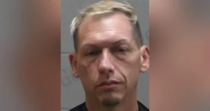 The image appears to be a mugshot of a man with short, light-colored hair, wearing a dark shirt.