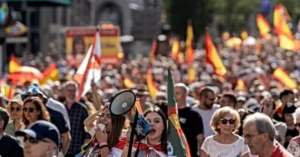 The image depicts a large crowd gathered outdoors, holding flags and participating in a public demonstration or rally.