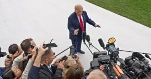 The image shows a public figure addressing a group of reporters and photographers, surrounded by microphones and cameras.