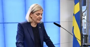 A woman in a dark suit stands at a podium with a microphone, delivering a speech in front of a Swedish flag.