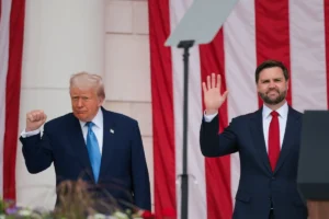 The image shows two men in suits standing in front of American flags, with one man raising his hand in a wave and the other making a fist gesture.