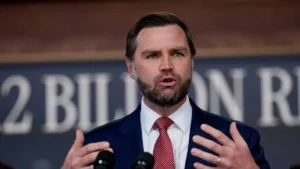 A man in a suit and tie is speaking at a press conference, with a backdrop that includes the text "2 BILLION."