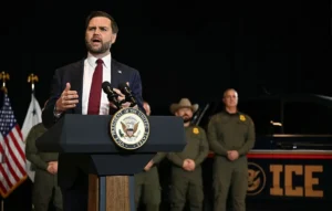 A man in a suit stands at a podium with the seal of the Vice President of the United States, addressing an audience with law enforcement personnel and an ICE vehicle in the background.