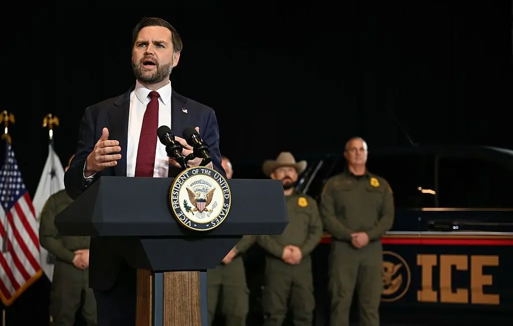 A man in a suit stands at a podium with the seal of the Vice President of the United States, addressing an audience with law enforcement personnel and an ICE vehicle in the background.
