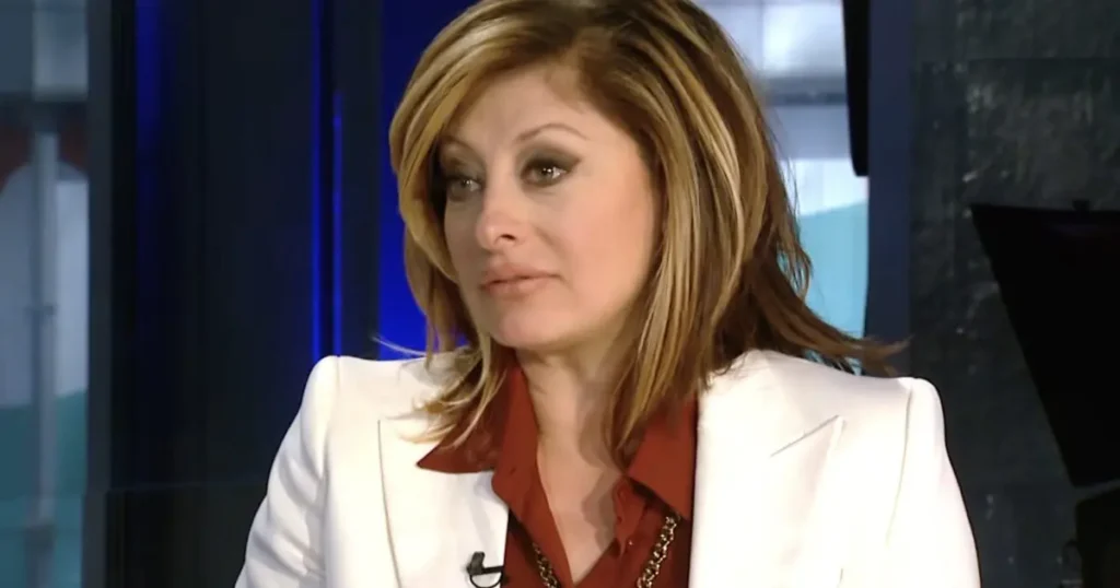 The image shows a woman in a professional setting, wearing a white blazer over a red shirt, likely during a news broadcast or interview.