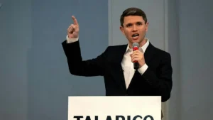A man in a suit is giving a speech at a podium with a microphone, gesturing with his right hand.