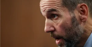 The image shows a close-up of a man with a beard and a light blue shirt, speaking with his mouth slightly open.