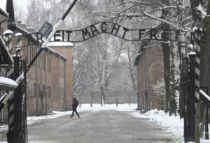 The image shows the entrance gate of Auschwitz-Birkenau, with the sign "Arbeit macht frei" (Work sets you free) partially covered in snow, symbolizing the historical and somber nature of the site.