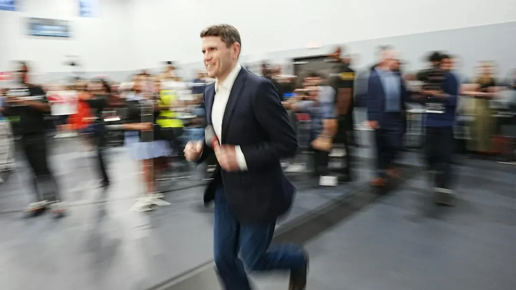 A man in a suit is running through a crowded indoor space, with blurred figures in the background.