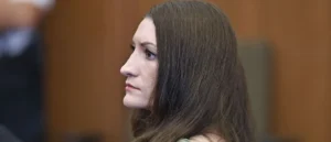 The image shows a person with long brown hair seated in what appears to be a formal setting, possibly a courtroom or a professional environment.