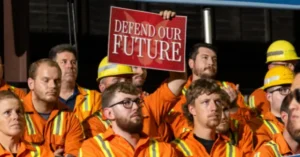 A group of workers in orange uniforms and hard hats holds a sign that reads "DEFEND OUR FUTURE."